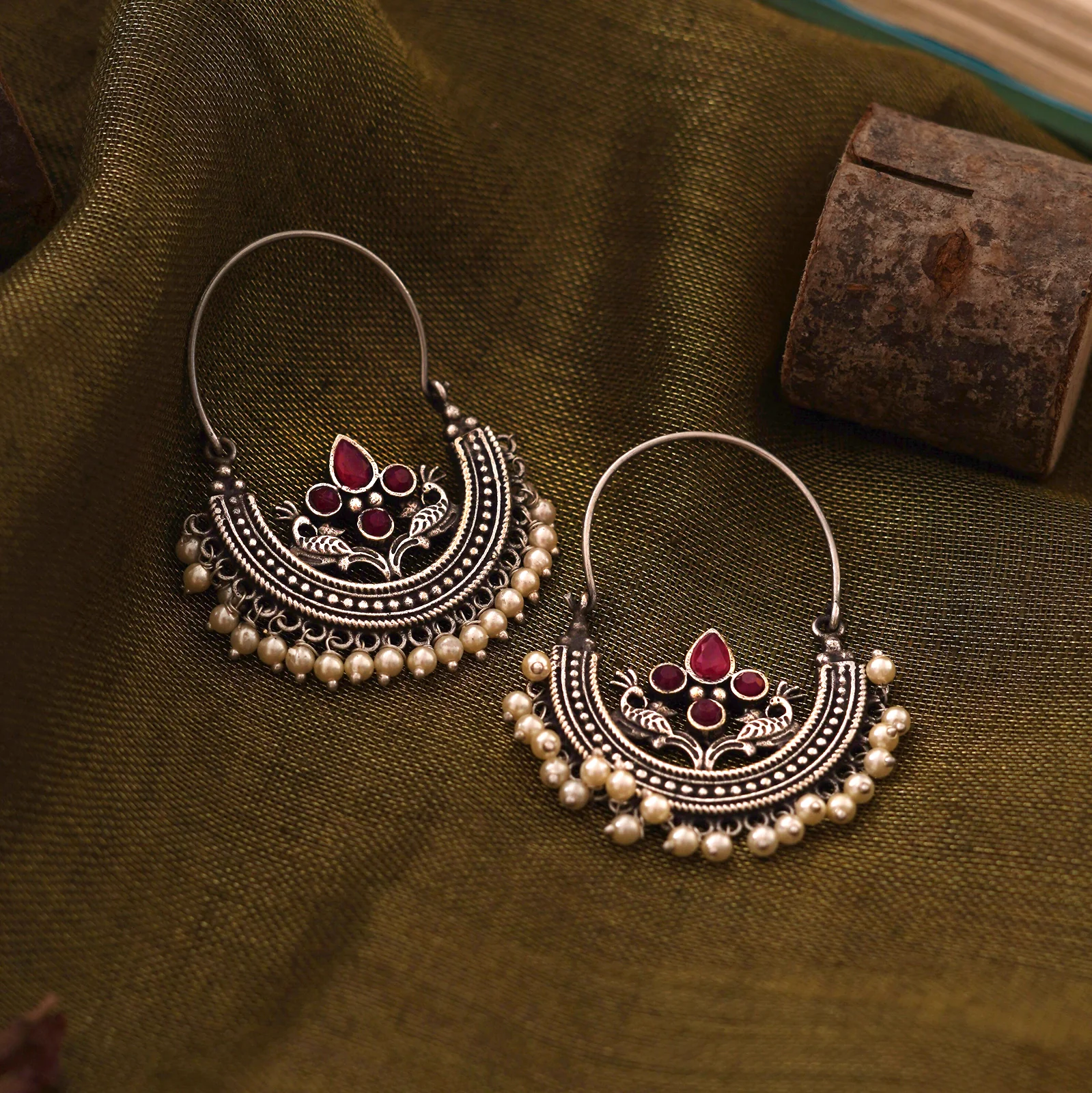Saara red stone oxidised hoop earrings with peacock motifs and faux pearl drops on olive fabric